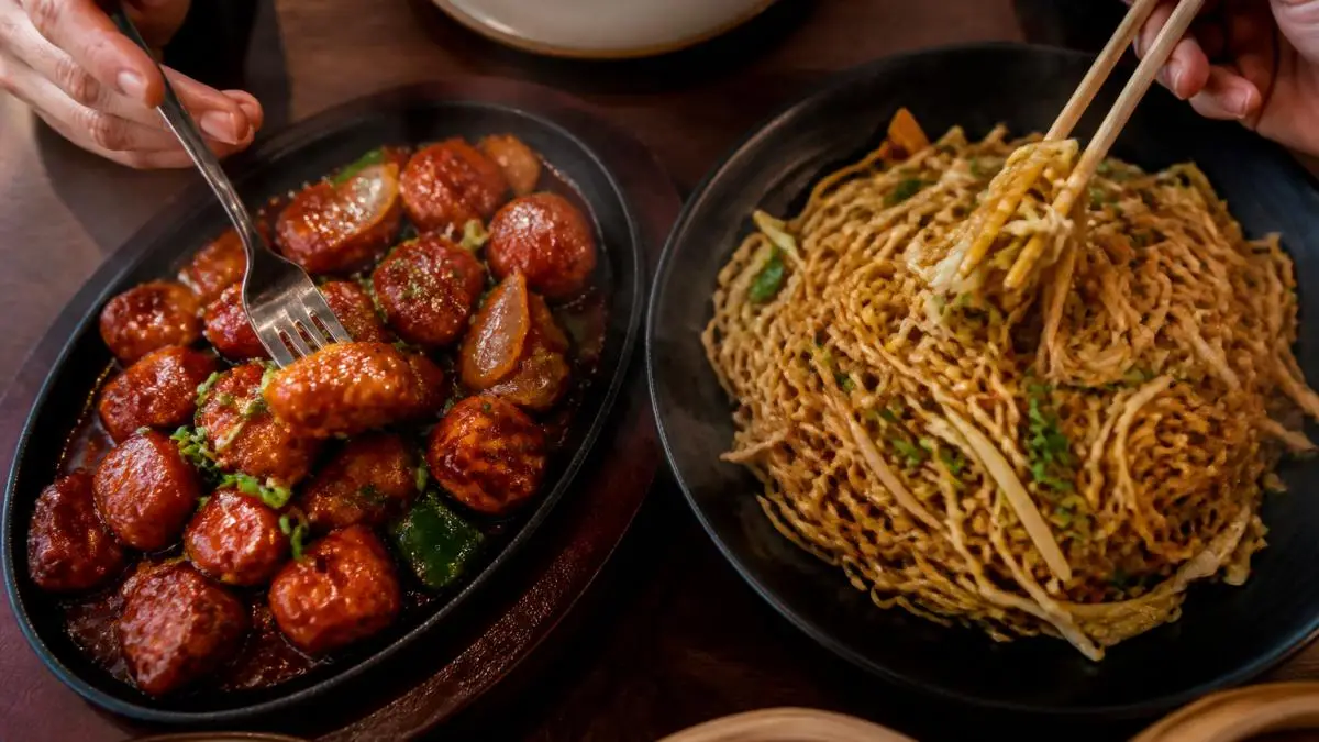 A close-up overhead shot of a sizzling plate of chilli paneer and hakka noodles on a warmly lit restaurant table, hands reaching in with chopsticks and a fork, steam rising from the food